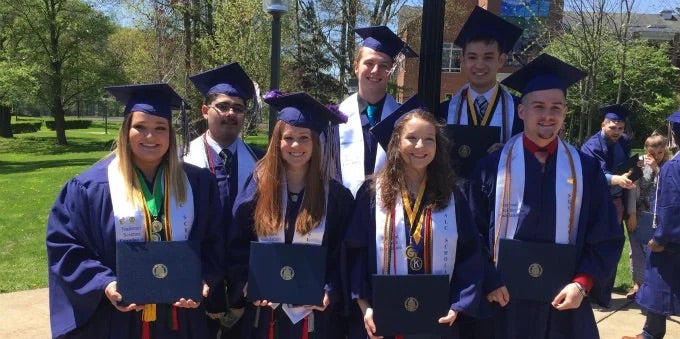 Seven student in graduation regalia holding diplomas.
